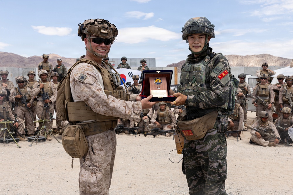 Two military personnel in the foreground are exchanging a commemorative plaque or award during what appears to be a joint military exercise or ceremony. One soldier wears US Marine Corps desert MARPAT camouflage, while the other wears a distinctive green camouflage uniform with a South Korean flag patch visible. A large group of assembled troops from both nations stands and sits in the background, with a South Korean flag visible.