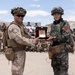 Two military personnel in the foreground are exchanging a commemorative plaque or award during what appears to be a joint military exercise or ceremony. One soldier wears US Marine Corps desert MARPAT camouflage, while the other wears a distinctive green camouflage uniform with a South Korean flag patch visible. A large group of assembled troops from both nations stands and sits in the background, with a South Korean flag visible.