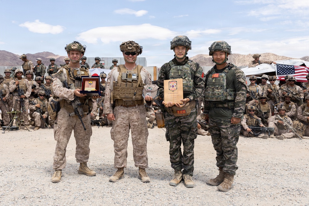 Four military personnel in full combat gear pose for a formal photo, with two holding plaques or awards. They appear to represent two different nations' armed forces, with US Marines in desert camouflage and soldiers in woodland/digital camouflage pattern uniforms. A large group of military personnel is assembled in the background near an American flag, suggesting a joint military exercise or ceremony.