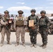 Four military personnel in full combat gear pose for a formal photo, with two holding plaques or awards. They appear to represent two different nations' armed forces, with US Marines in desert camouflage and soldiers in woodland/digital camouflage pattern uniforms. A large group of military personnel is assembled in the background near an American flag, suggesting a joint military exercise or ceremony.
