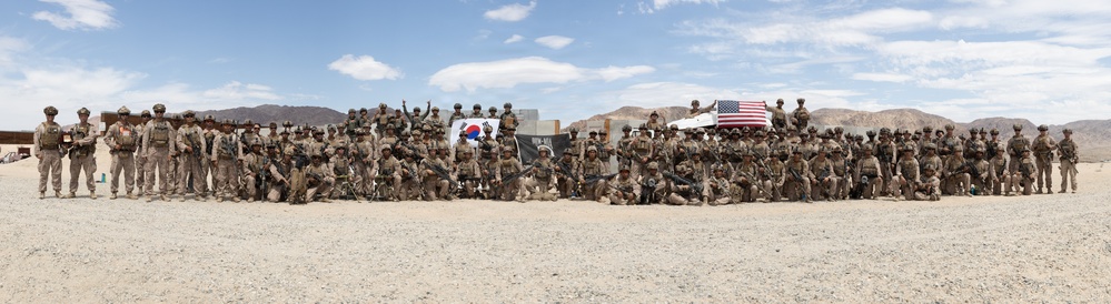 A large group of military personnel pose for a panoramic group photograph in an arid desert training environment. Two national flags are visible — the South Korean flag (Taegukgi) and the United States flag — suggesting a joint military exercise or training event. The setting appears to be a desert training base with mountainous terrain in the background.