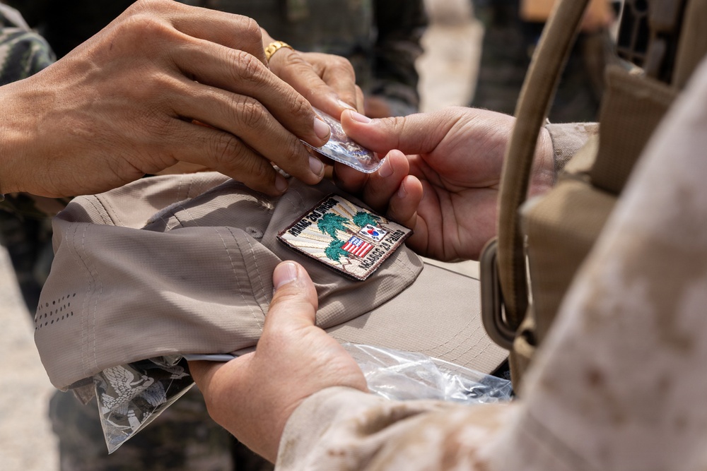 The image shows a close-up of two individuals exchanging or attaching a military patch onto a tan/khaki cap or hat. The patch features palm trees, what appears to be a South Korean flag, and an American shield emblem, suggesting a joint military exercise or commemorative exchange. One person appears to be in military uniform while the other is wearing civilian or different military attire.