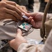 The image shows a close-up of two individuals exchanging or attaching a military patch onto a tan/khaki cap or hat. The patch features palm trees, what appears to be a South Korean flag, and an American shield emblem, suggesting a joint military exercise or commemorative exchange. One person appears to be in military uniform while the other is wearing civilian or different military attire.