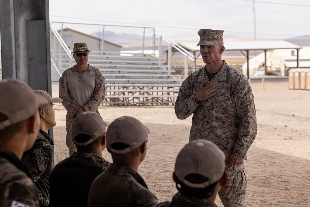 A US Marines officer, identifiable by the 'US MARINES' tape and rank insignia on his uniform, addresses a group of seated military personnel in what appears to be a desert training environment. The setting appears to be a military base or training facility, likely in an arid region based on the terrain and infrastructure visible in the background. The officer has his hand placed on his chest, suggesting a formal or emphatic moment during his address.