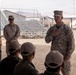 A US Marines officer, identifiable by the 'US MARINES' tape and rank insignia on his uniform, addresses a group of seated military personnel in what appears to be a desert training environment. The setting appears to be a military base or training facility, likely in an arid region based on the terrain and infrastructure visible in the background. The officer has his hand placed on his chest, suggesting a formal or emphatic moment during his address.