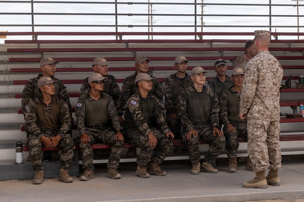 A group of approximately 11 South Korean military personnel in camouflage uniforms are seated on bleachers while a US Marine in desert MARPAT uniform addresses them. The South Korean soldiers are identifiable by the South Korean flag (Taegukgi) patches on their left shoulders. This appears to be a joint military training or briefing session, likely at a US military installation.