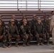 A group of approximately 11 South Korean military personnel in camouflage uniforms are seated on bleachers while a US Marine in desert MARPAT uniform addresses them. The South Korean soldiers are identifiable by the South Korean flag (Taegukgi) patches on their left shoulders. This appears to be a joint military training or briefing session, likely at a US military installation.