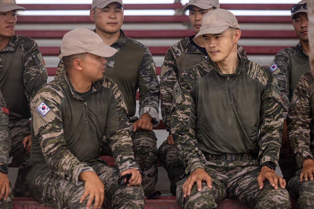 A group of military personnel wearing South Korean Army camouflage uniforms are seated on bleachers or stadium-style seating. Two soldiers in the foreground appear to be engaged in conversation, while others sit attentively in the background. The South Korean flag (Taegukgi) patches are clearly visible on their left sleeves.