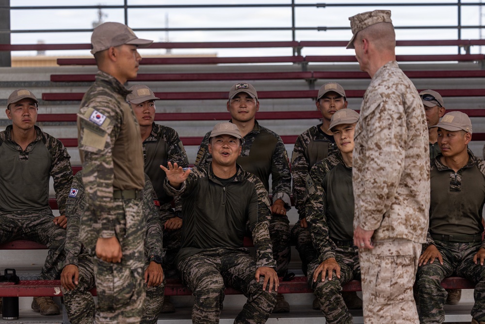 The image shows a group of South Korean military personnel seated in bleachers interacting with a U.S. Marine in desert MARPAT camouflage uniform. One South Korean soldier in the center appears to be speaking or asking a question with an open hand gesture, while a standing South Korean soldier and the U.S. Marine face each other in the foreground. This appears to be a joint military training or briefing session.