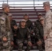 The image shows a group of South Korean military personnel seated in bleachers interacting with a U.S. Marine in desert MARPAT camouflage uniform. One South Korean soldier in the center appears to be speaking or asking a question with an open hand gesture, while a standing South Korean soldier and the U.S. Marine face each other in the foreground. This appears to be a joint military training or briefing session.