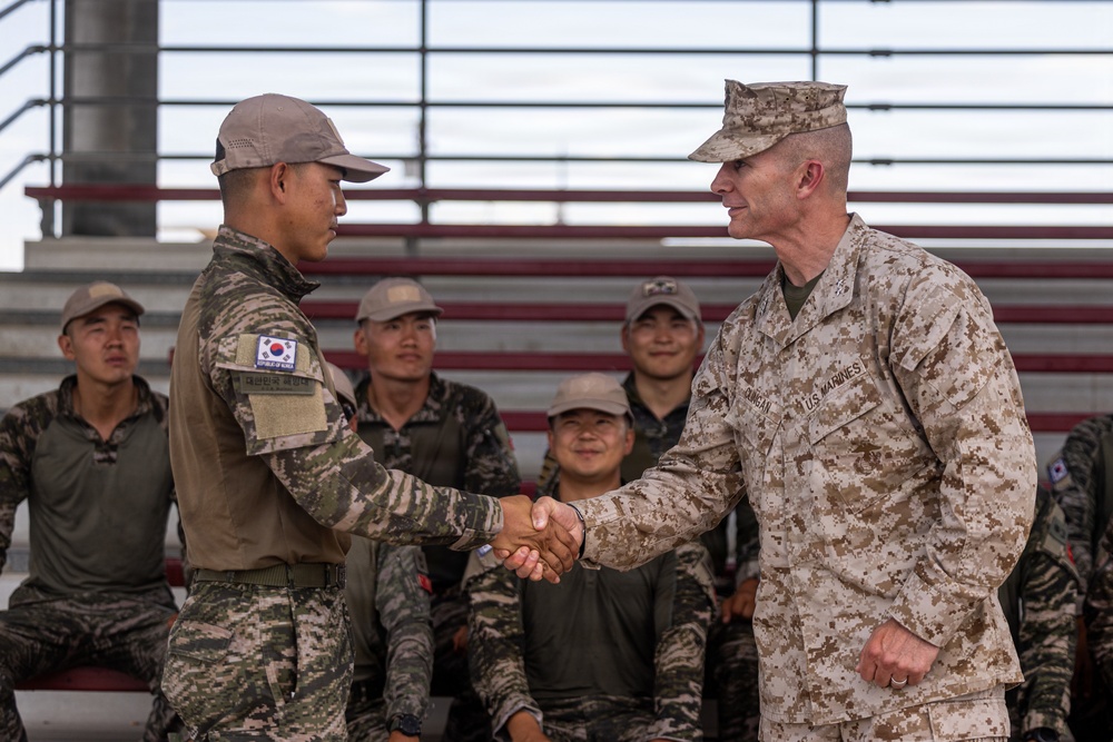 A U.S. Marine officer and a Republic of Korea (ROK) military soldier are shaking hands in what appears to be a formal or ceremonial exchange. Several ROK military personnel are seated in bleachers in the background, with at least one smiling. The setting suggests a joint military exercise or ceremony between U.S. and South Korean forces.