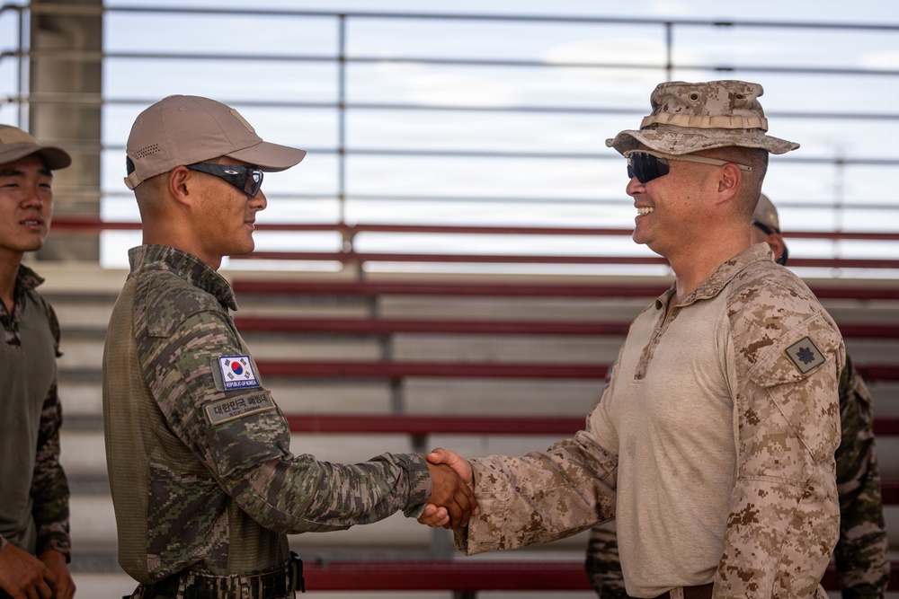 Two military personnel are shaking hands in what appears to be a formal greeting or ceremony. One individual wears a Republic of Korea (South Korean) military uniform with a visible Korean flag patch and Korean text, while the other wears US Marine Corps MARPAT desert camouflage with a cover (boonie hat) and a rank insignia consistent with a Major (gold oak leaf). The setting appears to be an outdoor military facility with bleachers visible in the background.