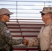 Two military personnel are shaking hands in what appears to be a formal greeting or ceremony. One individual wears a Republic of Korea (South Korean) military uniform with a visible Korean flag patch and Korean text, while the other wears US Marine Corps MARPAT desert camouflage with a cover (boonie hat) and a rank insignia consistent with a Major (gold oak leaf). The setting appears to be an outdoor military facility with bleachers visible in the background.