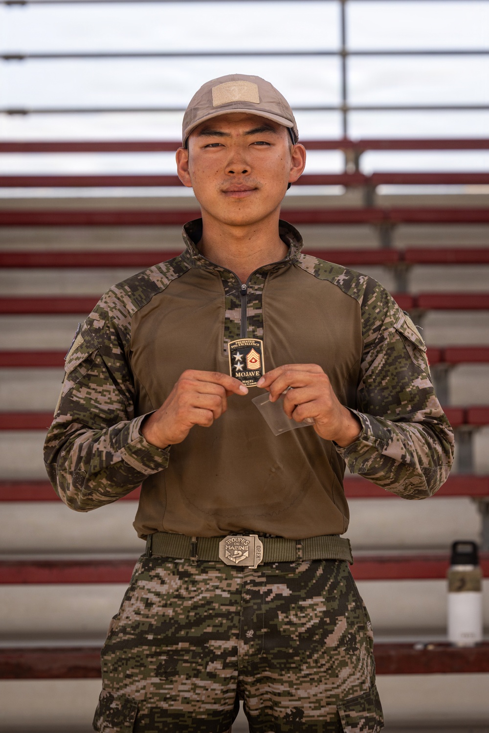 A young male soldier in camouflage uniform is posing in front of stadium bleachers, holding up what appears to be a challenge coin or award patch. The patch/coin reads 'MOJAVE' and includes text 'Presented for Excellence' along with a Master Sergeant rank insignia. He is wearing a mixed camouflage uniform with a belt buckle reading 'Troupes de Marine.'