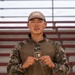 A young male soldier in camouflage uniform is posing in front of stadium bleachers, holding up what appears to be a challenge coin or award patch. The patch/coin reads 'MOJAVE' and includes text 'Presented for Excellence' along with a Master Sergeant rank insignia. He is wearing a mixed camouflage uniform with a belt buckle reading 'Troupes de Marine.'