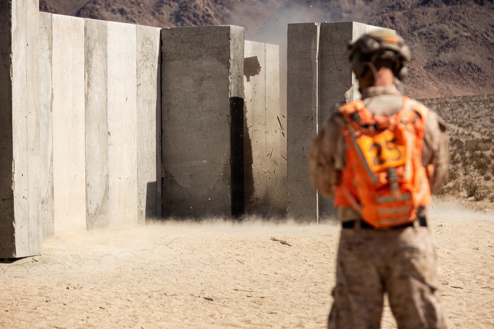 A military personnel wearing desert camouflage and an orange safety vest marked with the number '25' observes a controlled demolition or breaching exercise in a desert training environment. Concrete wall panels are being blown apart, with visible dust, debris, and blast effects. The setting appears to be a military training range with arid mountainous terrain in the background.
