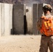 A military personnel wearing desert camouflage and an orange safety vest marked with the number '25' observes a controlled demolition or breaching exercise in a desert training environment. Concrete wall panels are being blown apart, with visible dust, debris, and blast effects. The setting appears to be a military training range with arid mountainous terrain in the background.