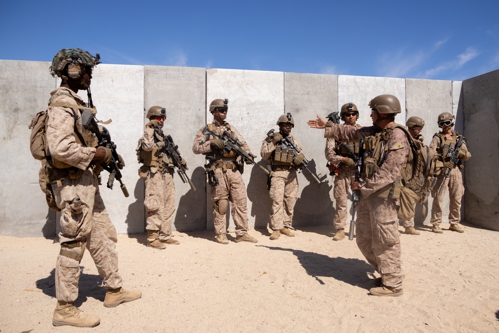 A group of approximately 8-10 military personnel in desert camouflage uniforms are assembled against a concrete barrier wall in an arid environment. One individual appears to be briefing or instructing the group, gesturing with an outstretched hand. The setting suggests a training exercise or pre-mission briefing at a desert military installation.