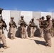 A group of approximately 8-10 military personnel in desert camouflage uniforms are assembled against a concrete barrier wall in an arid environment. One individual appears to be briefing or instructing the group, gesturing with an outstretched hand. The setting suggests a training exercise or pre-mission briefing at a desert military installation.