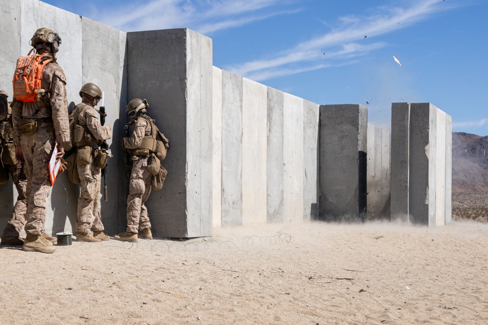 Military personnel in desert camouflage uniforms are taking cover against a concrete wall structure during what appears to be a controlled demolition or breaching exercise. An explosion or detonation is visibly occurring further along the concrete wall, with debris, dust, and fragments being ejected into the air. The setting appears to be a desert training range, consistent with facilities like those at Twentynine Palms or a similar U.S. military installation.