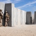Military personnel in desert camouflage uniforms are taking cover against a concrete wall structure during what appears to be a controlled demolition or breaching exercise. An explosion or detonation is visibly occurring further along the concrete wall, with debris, dust, and fragments being ejected into the air. The setting appears to be a desert training range, consistent with facilities like those at Twentynine Palms or a similar U.S. military installation.