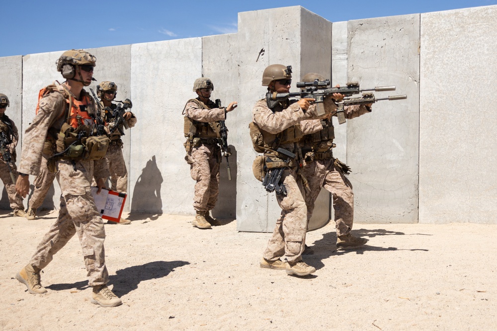A group of military personnel in desert camouflage uniforms are conducting what appears to be a tactical training exercise in an arid environment. They are maneuvering around large concrete barrier walls, with one soldier in a shooting stance aiming a rifle equipped with optics and a suppressor. One individual wearing an orange safety vest and carrying a clipboard appears to be an observer or evaluator for the exercise.