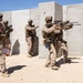 A group of military personnel in desert camouflage uniforms are conducting what appears to be a tactical training exercise in an arid environment. They are maneuvering around large concrete barrier walls, with one soldier in a shooting stance aiming a rifle equipped with optics and a suppressor. One individual wearing an orange safety vest and carrying a clipboard appears to be an observer or evaluator for the exercise.
