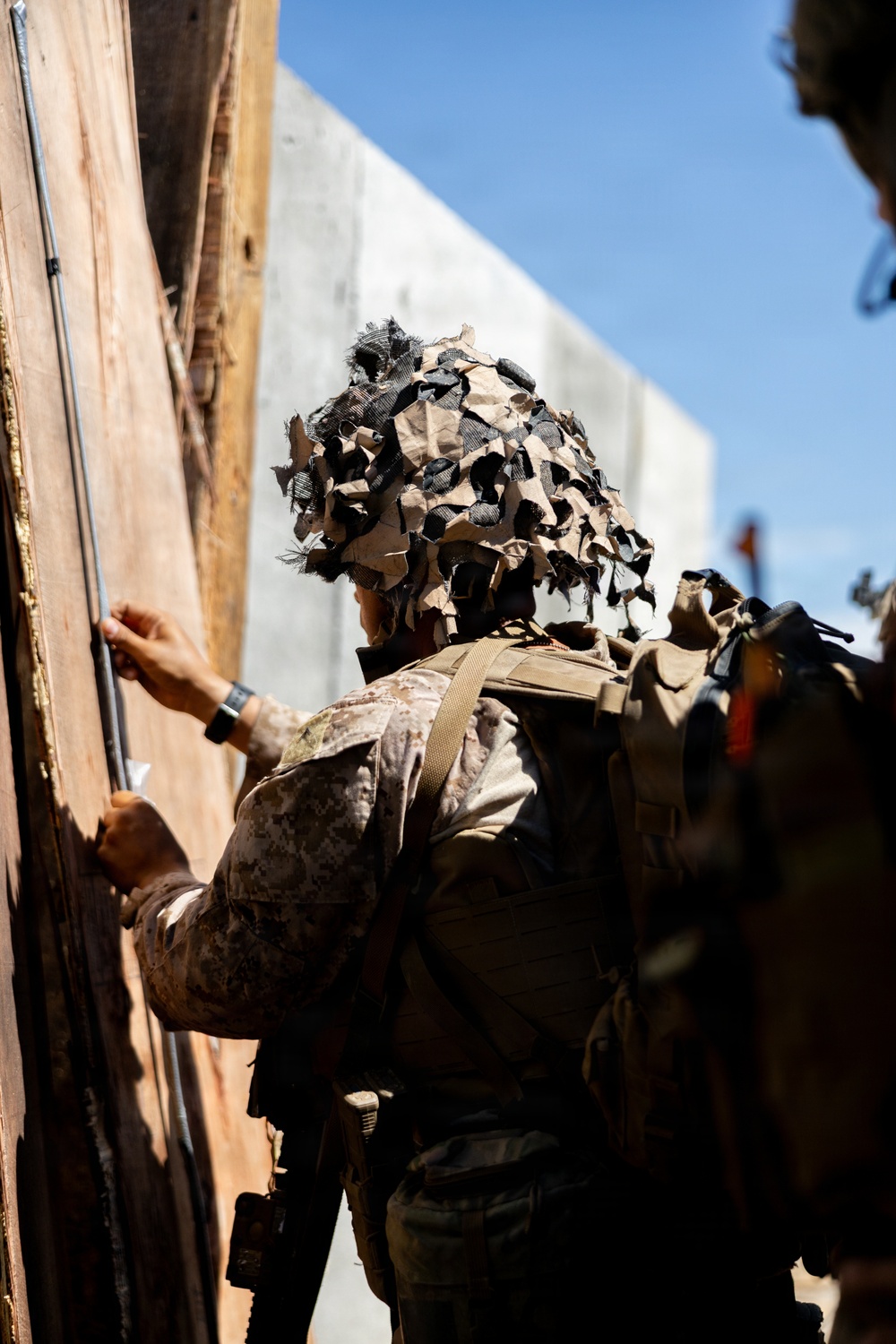 A military personnel wearing desert camouflage uniform and a camouflage ghillie-style head covering is working at a wooden door or wall structure. The individual appears to be placing or attaching something to the wooden surface, possibly a breaching charge or explosive device. At least one other similarly equipped personnel is visible in the background.