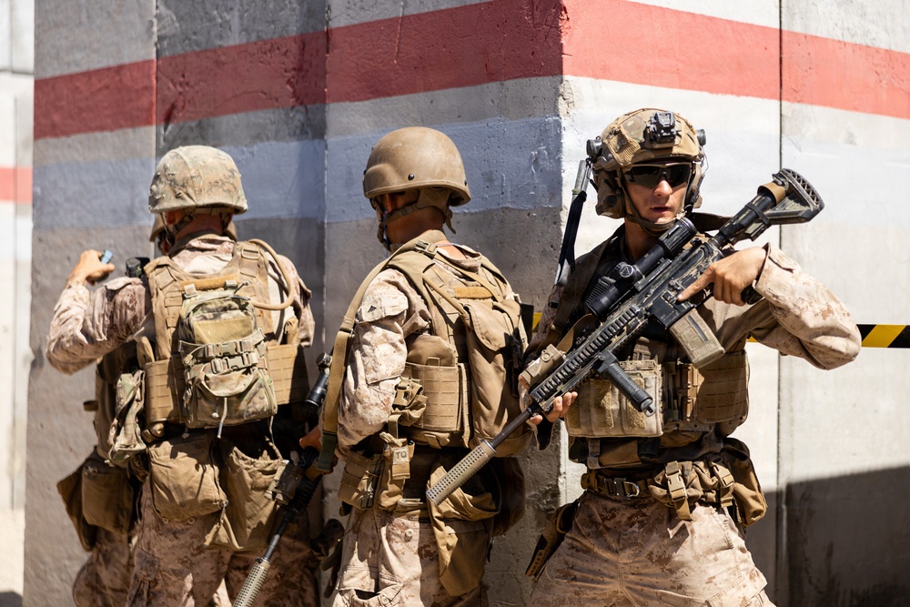 Three military personnel in desert camouflage uniforms are positioned against a concrete wall with red and white painted stripes. They appear to be conducting a tactical exercise or operation, with at least one soldier actively holding an assault rifle in a ready position. The setting suggests a training environment or urban operations scenario.