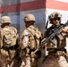 Three military personnel in desert camouflage uniforms are positioned against a concrete wall with red and white painted stripes. They appear to be conducting a tactical exercise or operation, with at least one soldier actively holding an assault rifle in a ready position. The setting suggests a training environment or urban operations scenario.