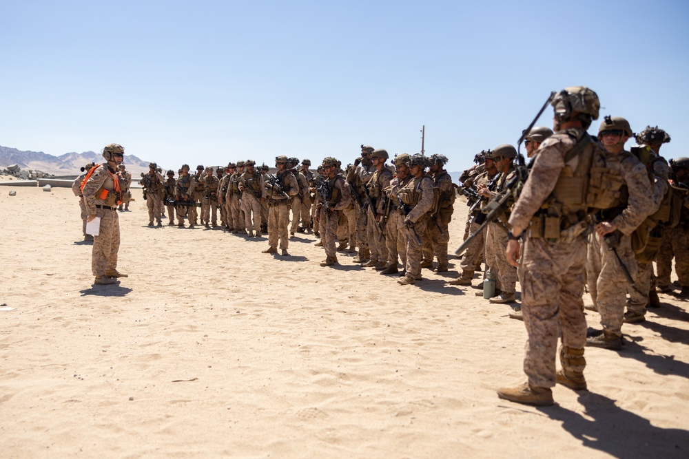 A large group of military personnel in desert camouflage uniforms are assembled in a sandy, arid environment. One individual wearing an orange safety vest stands facing the group, appearing to conduct a briefing or safety orientation. The setting suggests a training exercise or pre-mission briefing in a desert location.