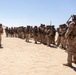 A large group of military personnel in desert camouflage uniforms are assembled in a sandy, arid environment. One individual wearing an orange safety vest stands facing the group, appearing to conduct a briefing or safety orientation. The setting suggests a training exercise or pre-mission briefing in a desert location.