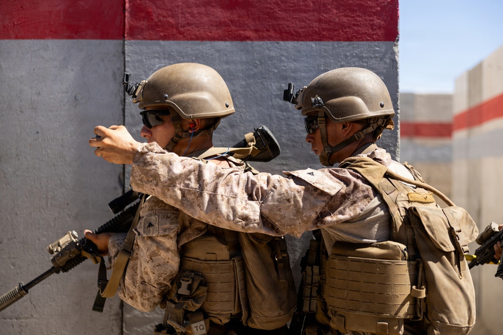 Two military personnel in full combat gear are positioned against a concrete wall, with one soldier appearing to point or gesture toward a direction. Both are equipped with tactical vests, helmets, weapons, and protective eyewear, suggesting a training exercise or operational scenario. The wall behind them features red and gray painted sections, consistent with a military training facility.