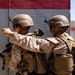 Two military personnel in full combat gear are positioned against a concrete wall, with one soldier appearing to point or gesture toward a direction. Both are equipped with tactical vests, helmets, weapons, and protective eyewear, suggesting a training exercise or operational scenario. The wall behind them features red and gray painted sections, consistent with a military training facility.