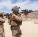 The image shows multiple military personnel in desert camouflage uniforms conducting what appears to be a live-fire training exercise in an arid desert environment. The soldiers are equipped with full tactical gear including body armor, helmets, and backpacks, and are aiming rifles downrange. The setting appears to be a designated military training range based on the organized firing line formation.