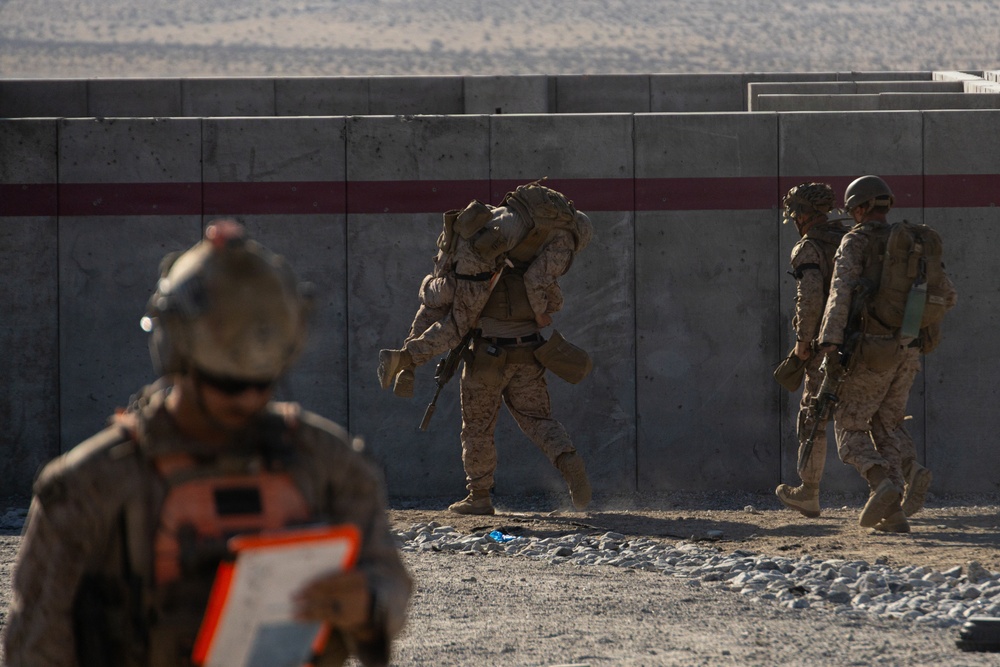 The image shows military personnel in desert camouflage uniforms conducting what appears to be a training exercise near a concrete barrier wall in an arid environment. One soldier in the center is carrying a heavy load or simulated casualty on their back, while others move alongside the wall. A fourth individual in the foreground, slightly out of focus, appears to be an observer or evaluator holding a clipboard with an orange backing.