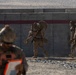 The image shows military personnel in desert camouflage uniforms conducting what appears to be a training exercise near a concrete barrier wall in an arid environment. One soldier in the center is carrying a heavy load or simulated casualty on their back, while others move alongside the wall. A fourth individual in the foreground, slightly out of focus, appears to be an observer or evaluator holding a clipboard with an orange backing.