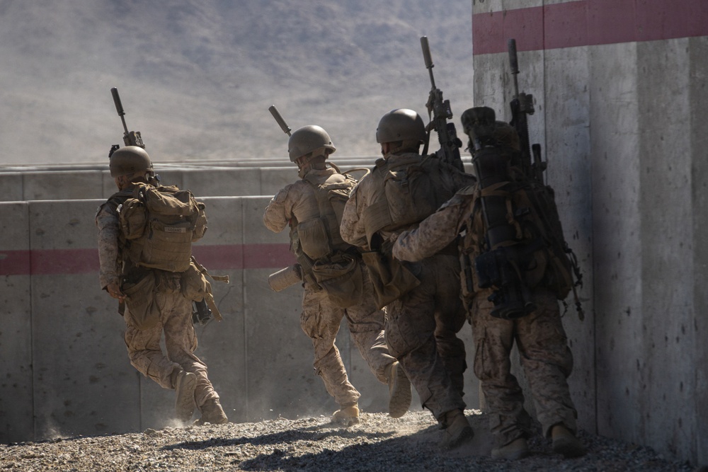 Four military personnel in desert camouflage uniforms are conducting a tactical movement exercise near a concrete barrier wall. The soldiers are carrying rifles and full combat gear, appearing to stack up against a wall in a breach or entry formation. The setting appears to be a training facility or urban operations range based on the structured concrete barriers with red stripe markings.