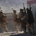 Four military personnel in desert camouflage uniforms are conducting a tactical movement exercise near a concrete barrier wall. The soldiers are carrying rifles and full combat gear, appearing to stack up against a wall in a breach or entry formation. The setting appears to be a training facility or urban operations range based on the structured concrete barriers with red stripe markings.