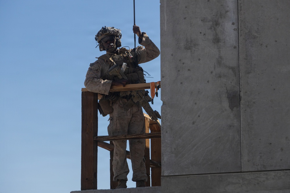 A fully equipped military service member in desert camouflage (MARPAT pattern) stands on an elevated wooden platform adjacent to a concrete structure, gripping a rope above their head. The individual is wearing full tactical gear including a helmet with communications equipment, body armor, and carrying a rifle. This appears to be a training exercise, likely involving rappelling or fast-rope insertion/extraction techniques.