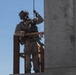 A fully equipped military service member in desert camouflage (MARPAT pattern) stands on an elevated wooden platform adjacent to a concrete structure, gripping a rope above their head. The individual is wearing full tactical gear including a helmet with communications equipment, body armor, and carrying a rifle. This appears to be a training exercise, likely involving rappelling or fast-rope insertion/extraction techniques.