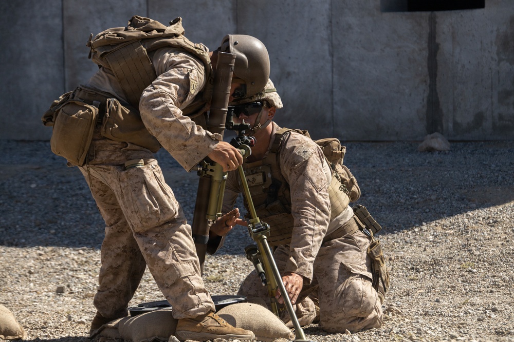 Two military personnel in desert camouflage uniforms are working together to set up or adjust a mortar system on a gravel surface. Both are wearing full combat gear including body armor, tactical vests, and backpacks. The setting appears to be a training area or forward operating base, indicated by the concrete barrier wall in the background.