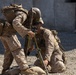 Two military personnel in desert camouflage uniforms are working together to set up or adjust a mortar system on a gravel surface. Both are wearing full combat gear including body armor, tactical vests, and backpacks. The setting appears to be a training area or forward operating base, indicated by the concrete barrier wall in the background.
