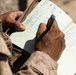 A person wearing military camouflage (MARPAT pattern) uniform is writing in a ruled notebook with a blue pen. The notebook contains handwritten numerical data organized in columns with headers including 'DEF' and 'CH'. The setting appears to be outdoors in a field or operational environment.