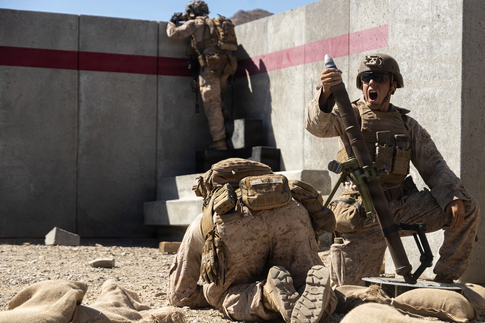 The image shows U.S. Marines conducting what appears to be a military training exercise at a concrete training facility. One Marine is holding a mortar round aloft while shouting, positioned next to a mortar tube on a bipod, while a second Marine is prone on the ground and a third climbs concrete steps in the background. The arid, desert-like environment and training infrastructure suggest a stateside or overseas military training range.