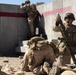 The image shows U.S. Marines conducting what appears to be a military training exercise at a concrete training facility. One Marine is holding a mortar round aloft while shouting, positioned next to a mortar tube on a bipod, while a second Marine is prone on the ground and a third climbs concrete steps in the background. The arid, desert-like environment and training infrastructure suggest a stateside or overseas military training range.