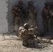 The image shows a group of military personnel in desert camouflage uniforms conducting what appears to be a tactical training exercise or combat operation near a concrete wall. Two soldiers are in a prone/kneeling position operating a belt-fed machine gun in the foreground, while several others stand against the wall in a stack formation. The environment is arid and dusty, consistent with a desert or semi-arid training environment.