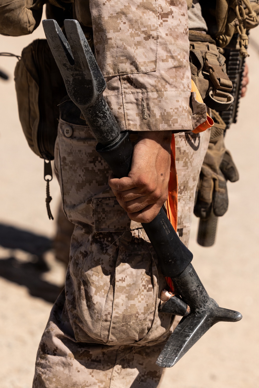 A close-up image of a military personnel wearing USMC MARPAT desert camouflage uniform holding a Halligan bar (breaching tool) with a black handle and orange strap. The individual is equipped with tactical gear including a backpack and what appears to be a firearm visible in the background. The setting appears to be an arid, sandy outdoor environment.