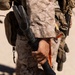 A close-up image of a military personnel wearing USMC MARPAT desert camouflage uniform holding a Halligan bar (breaching tool) with a black handle and orange strap. The individual is equipped with tactical gear including a backpack and what appears to be a firearm visible in the background. The setting appears to be an arid, sandy outdoor environment.