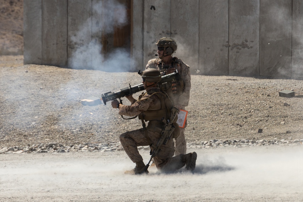 Two military personnel in desert camouflage uniforms are firing a shoulder-launched rocket or recoilless weapon system, with visible backblast smoke and dust cloud generated by the discharge. One soldier is in a kneeling position operating the weapon while a second stands directly behind, both wearing combat helmets and tactical gear. The setting appears to be a training range or arid operational environment with a concrete barrier wall in the background.